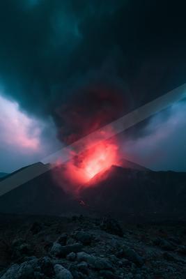 A powerful volcanic eruption at night, with bright red glowing lava and a massive plume of dark smoke rising into the sky above a rocky volcanic landscape.