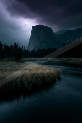 Moody landscape with a large granite mountain under dark storm clouds, a flowing river in the foreground, and a forest of conifer trees.