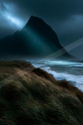 A dramatic stormy coastal landscape featuring a large dark rocky mountain under heavy clouds, with turbulent ocean waves crashing on the shore and windswept gra