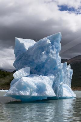 A massive, jagged blue and white iceberg floats in a calm lake with dark mountains and a cloudy sky in the background.
