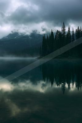 A serene lake surrounded by evergreen trees reflects a cloudy sky, creating a mirror-like surface in the misty atmosphere.