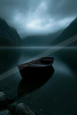 A dark wooden rowboat floats on the calm, reflective surface of a misty mountain lake, surrounded by steep forested slopes under a foggy, overcast sky.