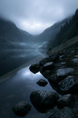 A calm mountain lake reflects the foggy, overcast sky and steep forested slopes, with large dark rocks lining the misty shoreline in a serene, atmospheric scene