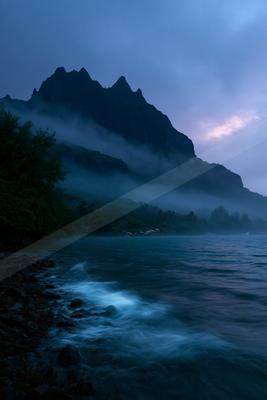 A dramatic coastal landscape at dusk featuring a large jagged mountain shrouded in thick mist, a rocky shoreline with glowing waves, and dark tropical vegetatio