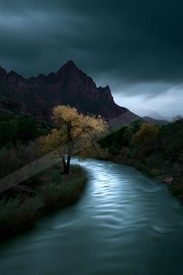 A smooth flowing river winds through a rugged landscape with a lone yellow-leafed tree on the bank, backed by towering red rock mountains beneath a dark, overca