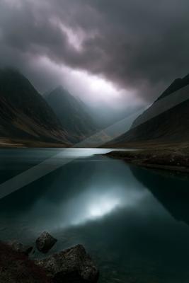 A serene alpine lake reflects the dramatic overcast sky and surrounding rugged mountains in a moody, atmospheric landscape.