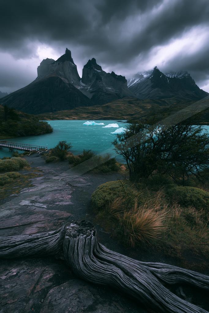 A moody mountain range looms over a serene turquoise lake under dark, brooding clouds. A wooden path leads through the foreground, flanked by shrubs and fallen