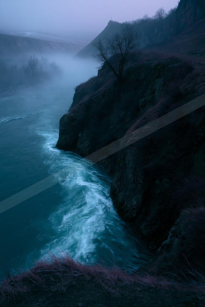 A powerful river with white rapids flows through a steep rocky canyon shrouded in thick blue mist, with a lone bare tree on the cliff edge under an overcast sky