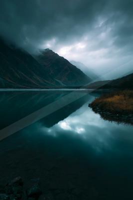 A calm mountain lake reflects the steep, cloud-covered peaks and overcast sky in moody blue tones, with rocky foreground and autumnal vegetation on the shore.