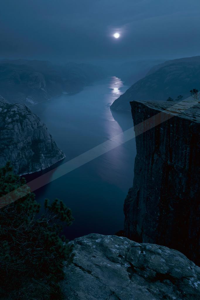 A dramatic night view of a deep fjord with steep rocky cliffs under a bright full moon, its light reflecting on the calm water surface, surrounded by misty moun