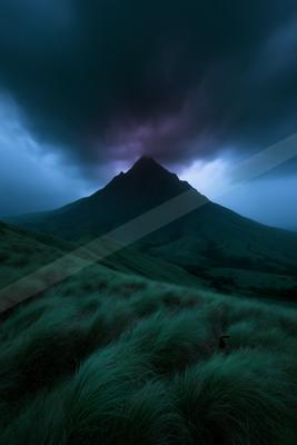 A solitary mountain peak rises against a dramatic, dark stormy sky filled with deep purple and blue clouds, with lush green grassy hills in the foreground.