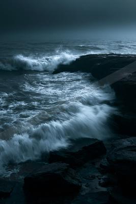 Powerful white waves crashing against dark jagged rocks under a moody, overcast sky, creating a dramatic seascape with motion blur in the turbulent water.