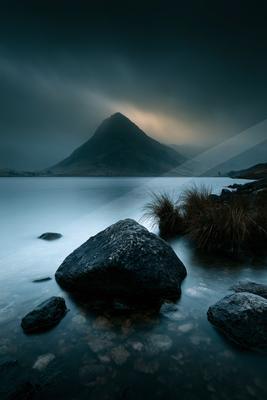 A large dark rock sits in the foreground of a calm lake, with a towering mountain partially shrouded in mist and dark storm clouds overhead.