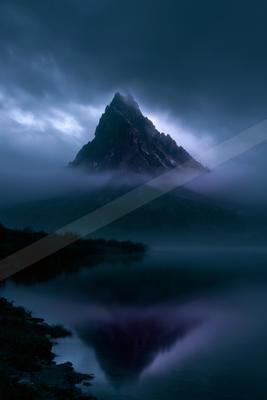 A dramatic, jagged mountain peak rises through thick fog and low clouds, perfectly reflected in the still surface of a foreground lake under a dark, moody sky.