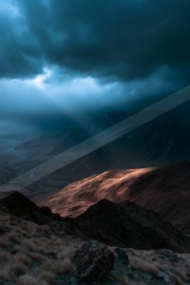 Dramatic crepuscular rays pierce through dark storm clouds, illuminating a vast mountain valley with a mix of cool blue tones and warm golden light on the slope