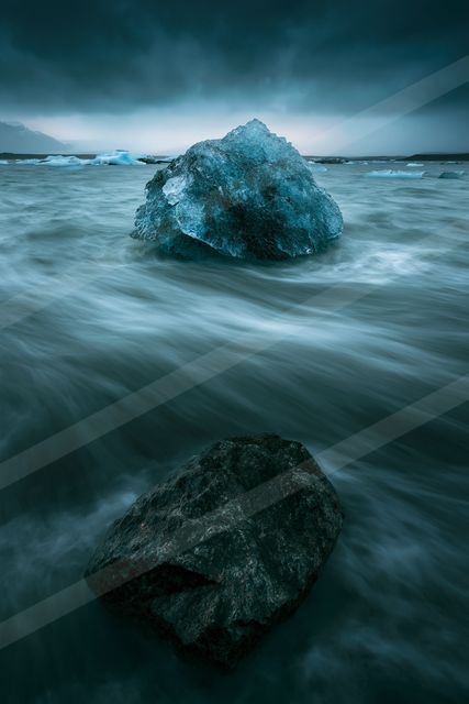 A large iceberg floats serenely in calm, turquoise waters under a moody sky. A dark rock is partially submerged in the foreground.