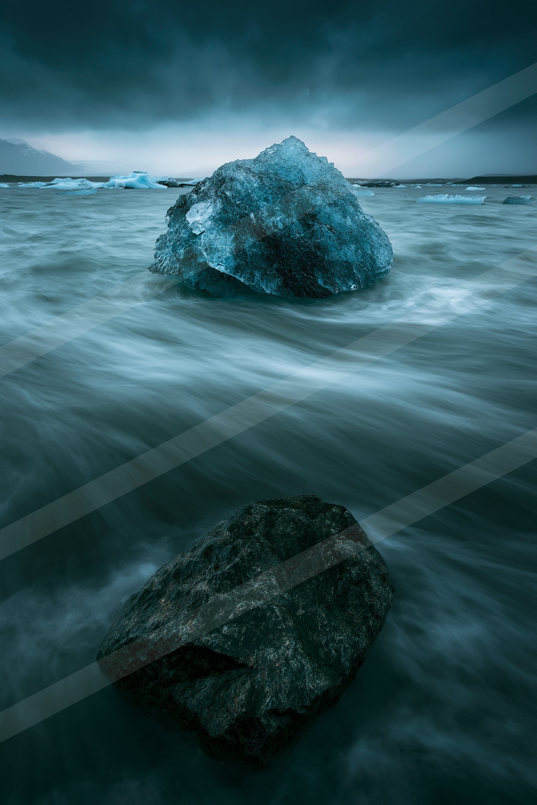 A large iceberg floats serenely in calm, turquoise waters under a moody sky. A dark rock is partially submerged in the foreground.
