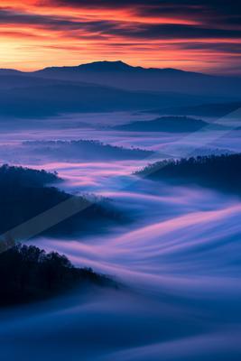A stunning landscape featuring a mountain range silhouetted against a vibrant sunset sky, with rolling fog blanketing the valleys below.