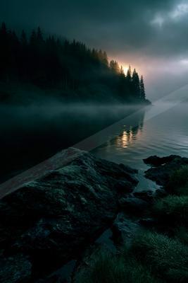 A serene misty lake at dawn with a dense coniferous forest on the hillside, large rocks and grass in the foreground, and a faint sun breaking through the clouds