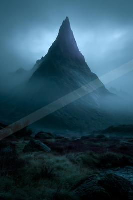 A sharp rocky mountain peak rises through thick fog and low clouds in a moody, atmospheric landscape with rocky foreground and sparse vegetation.