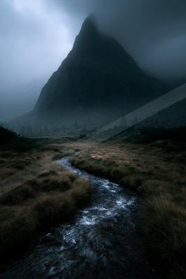 A rushing stream flows through a grassy valley toward a large, fog-shrouded mountain under a moody, overcast sky.