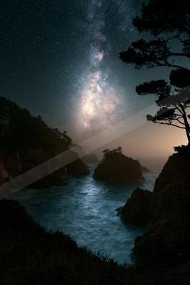 A dramatic night scene featuring the Milky Way galaxy arching across a starry sky above a rugged rocky coastline, with silhouetted pine trees and glowing ocean 