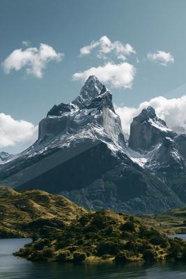 A majestic snow-covered mountain range rises against a clear blue sky with scattered clouds, reflected in the calm waters of a serene lake below.