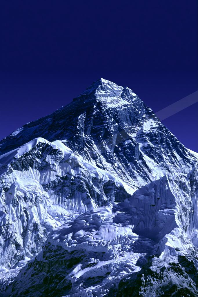 A dramatic, snow-covered mountain peak rises sharply against a deep blue sky, showcasing rugged icy ridges and steep rock faces.