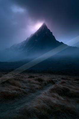 A sharp, rocky mountain peak rises dramatically through thick, dark storm clouds illuminated by a purple light from behind, with a misty foreground of dry grass