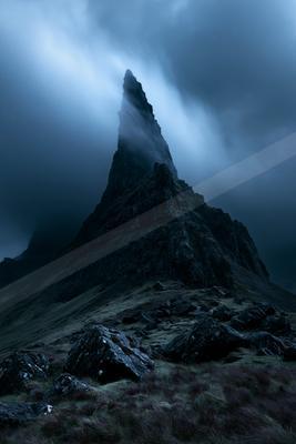 A sharp rocky mountain peak rises dramatically into thick swirling mist and dark storm clouds, with a foreground of rugged boulders and purple-tinged grass.