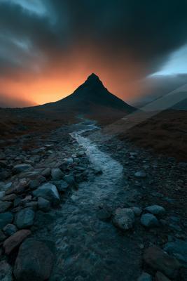 A towering mountain peak stands silhouetted against a vibrant sunset sky, with a rocky path leading towards it through rugged terrain.