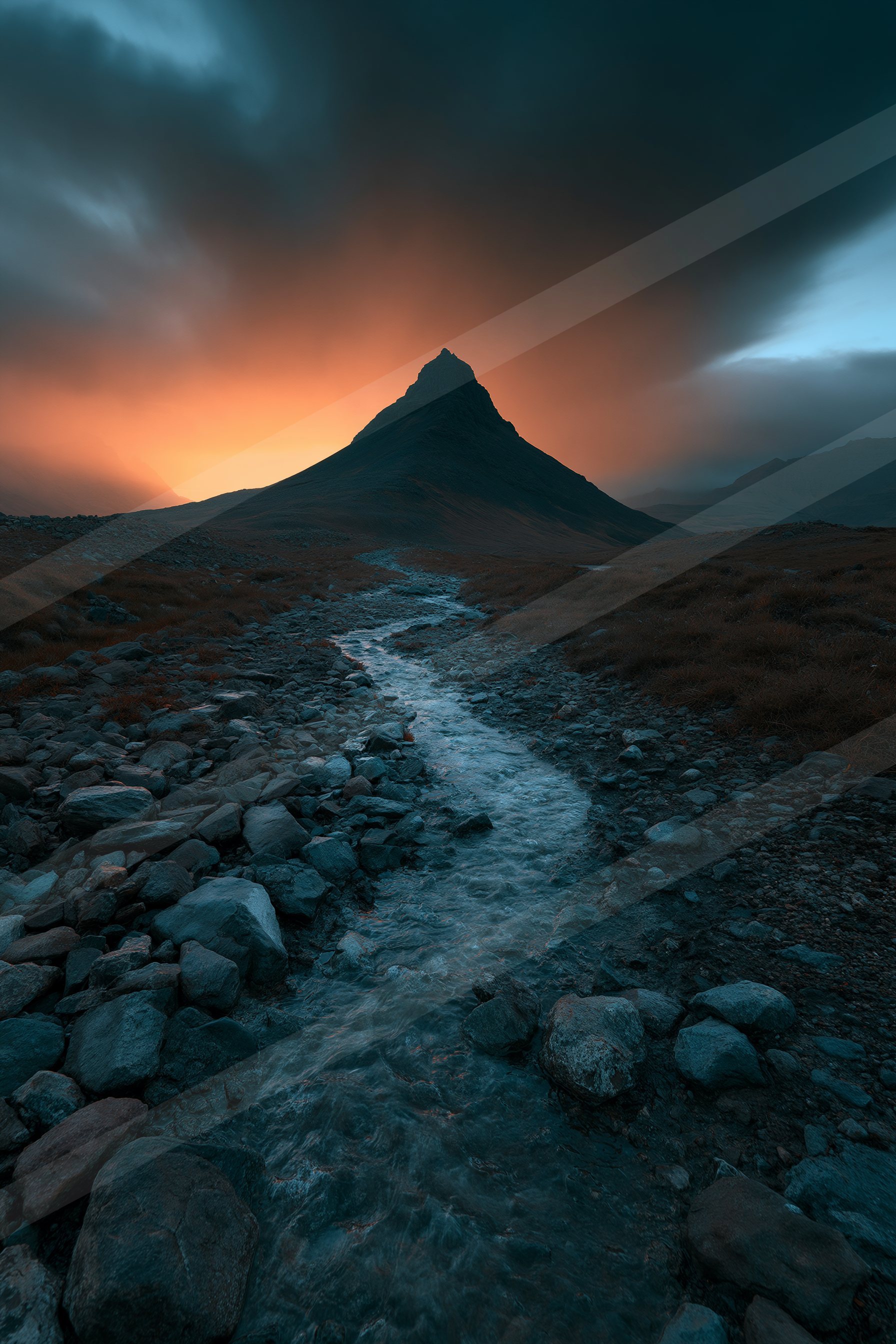 A towering mountain peak stands silhouetted against a vibrant sunset sky, with a rocky path leading towards it through rugged terrain.
