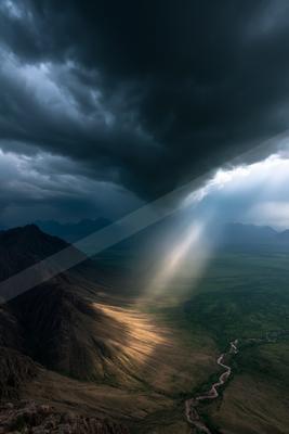 Moody landscape with dark storm clouds and crepuscular rays illuminating a vast mountain valley and winding river.
