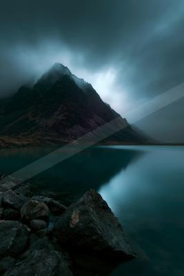 A rugged mountain peak partially shrouded in thick fog rises above a perfectly still lake that mirrors the moody blue and teal tones, with dark rocks in the for