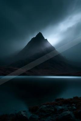 A sharp, dark mountain peak rises through thick fog above a perfectly still lake under a brooding, cloudy sky, with rocky foreground and reddish moorland.