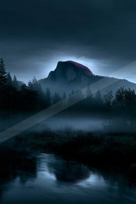 A large rocky mountain peak rises above a dense coniferous forest shrouded in thick fog, with a flowing river in the foreground under a dark, moody sky.