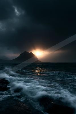 Turbulent dark ocean waves crash in the foreground under a brooding stormy sky, with a silhouetted mountain range and a bright sun breaking through the clouds i