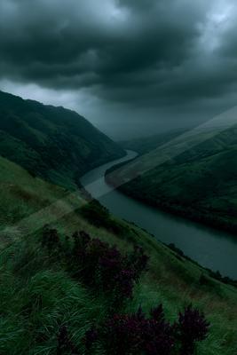 A dramatic landscape featuring a winding river flowing through a lush green valley under heavy dark storm clouds, with purple heather in the foreground on a gra