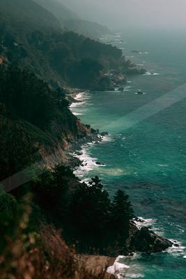 Aerial view of a rugged coastline with steep forested cliffs meeting turquoise ocean waters under heavy fog.