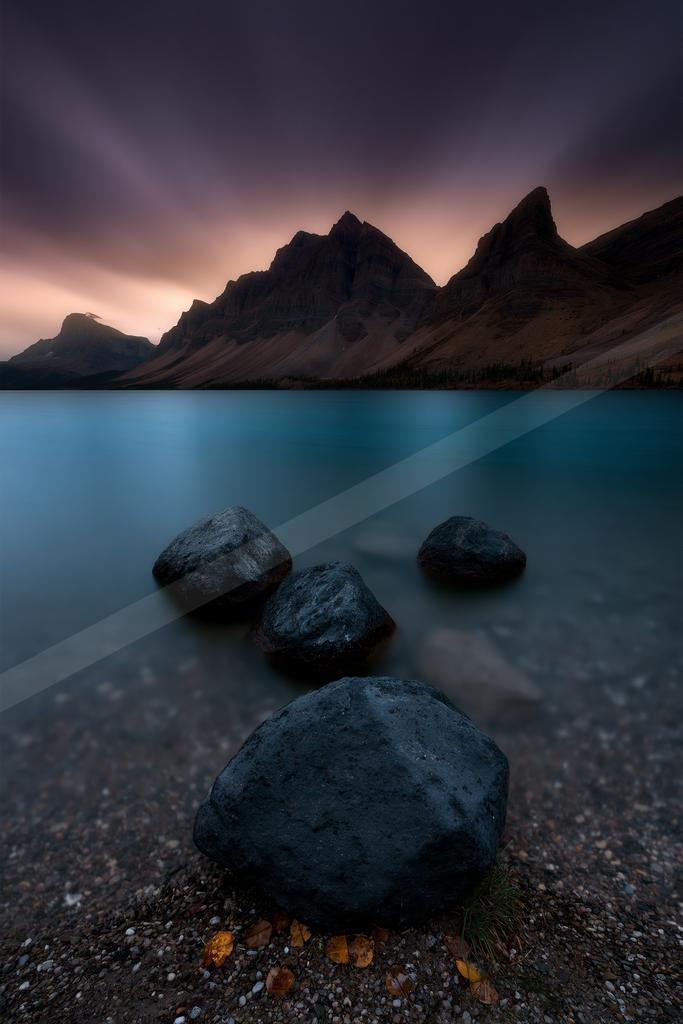 Large dark rocks rest on a pebbly shore in the foreground of a calm turquoise mountain lake, with a dramatic jagged mountain range silhouetted against a purple