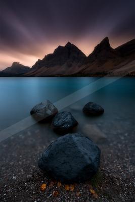 Large dark rocks rest on a pebbly shore in the foreground of a calm turquoise mountain lake, with a dramatic jagged mountain range silhouetted against a purple 
