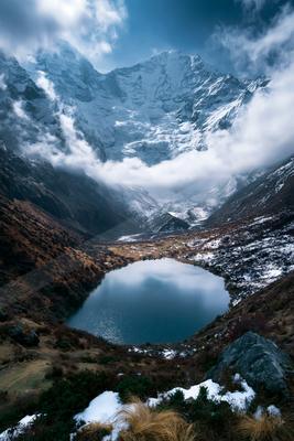 A serene mountain lake nestled between rugged, snow-covered peaks with clouds swirling around the summit. The foreground features a mix of dry grasses and patch