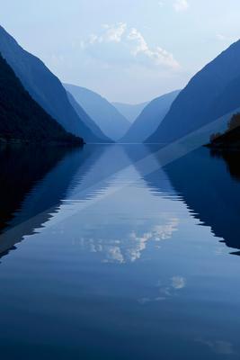 Calm fjord water perfectly reflecting steep forested mountains and a blue sky with clouds in a symmetrical composition.