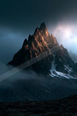 A rugged, snow-dusted mountain peak rises sharply into a dark, cloudy sky, illuminated by a warm light breaking through the mist.