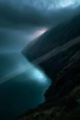 A dramatic moody landscape of a deep blue fjord winding between steep dark mountains under heavy storm clouds, with a subtle glow of light breaking through the 