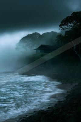 A dark wooden cabin sits on a foggy, tree-covered cliff above a turbulent ocean with waves crashing on the rocky shore under a stormy sky.