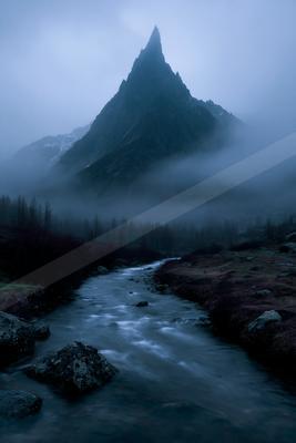 A dramatic, jagged mountain peak rises through thick fog above a flowing river in a misty wilderness landscape.