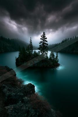 A small rocky island covered in tall evergreen trees sits in the middle of a turquoise mountain lake, surrounded by forested hills under a dark, cloudy sky with