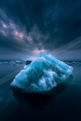 A massive, glowing blue iceberg floats in the calm, dark waters of an arctic sea under a dramatic, cloudy twilight sky.