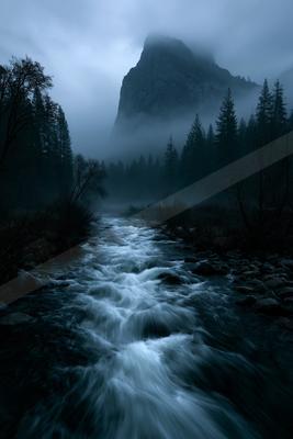 A fast-flowing river rushes over rocks through a misty, dark forest with tall conifer trees lining the banks, leading toward a large rocky mountain peak shroude